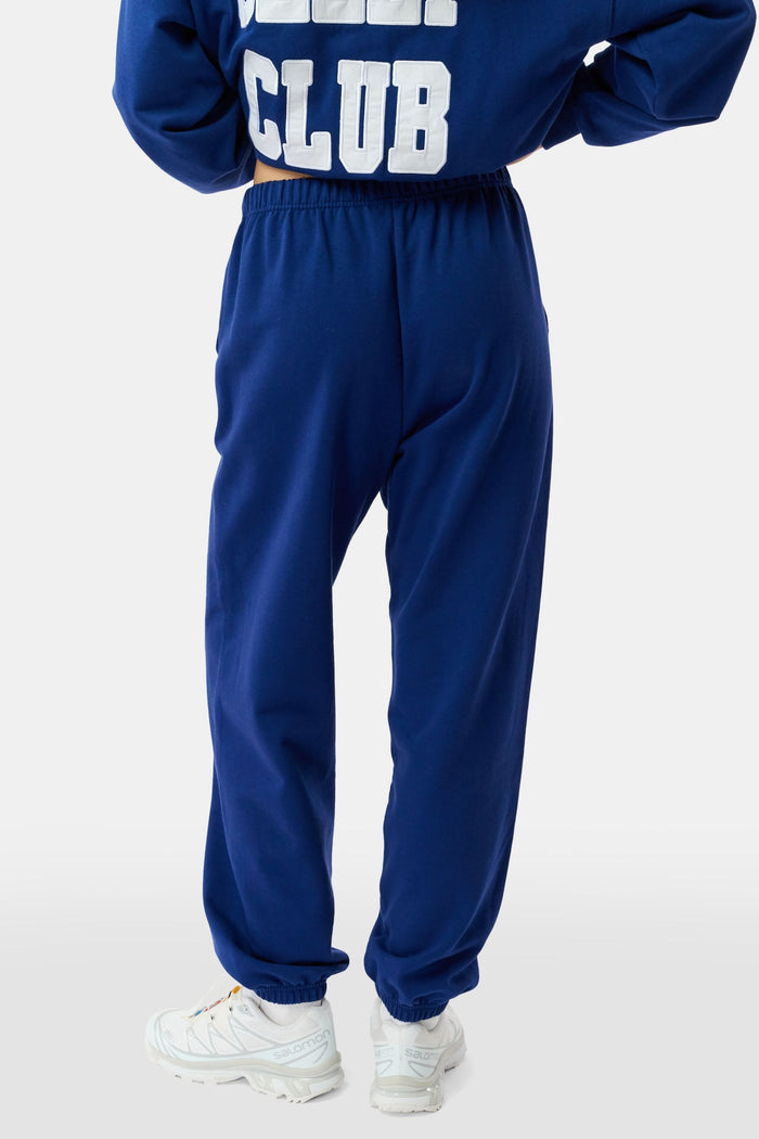 A young woman faces forward in a matching blue hoodie and Roller Rabbit Sleep Club Bea Sweatpants with "SLEEP CLUB" printed in white, paired with white sneakers. The hoodie has a small white graphic.