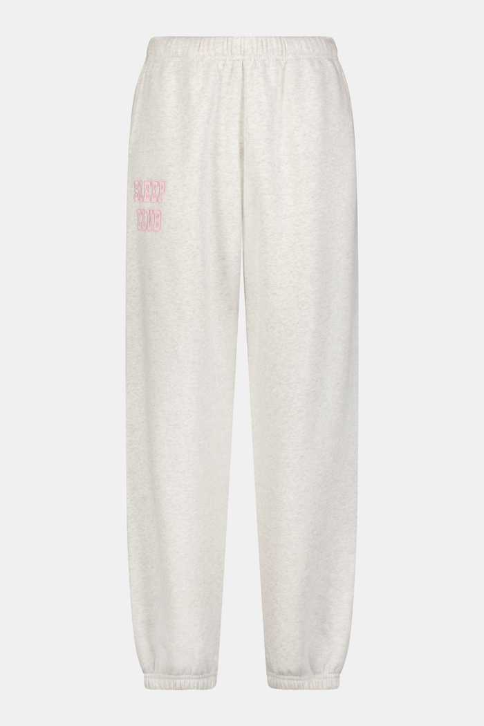 A woman stands against a white background wearing light gray Roller Rabbit Sleep Club Bea Sweatpants and a matching sweatshirt with "SLEEP CLUB" in pink, styled with pink and black Adidas sneakers.