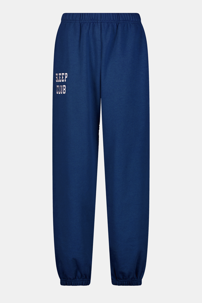 A young woman faces forward in a matching blue hoodie and Roller Rabbit Sleep Club Bea Sweatpants with "SLEEP CLUB" printed in white, paired with white sneakers. The hoodie has a small white graphic.