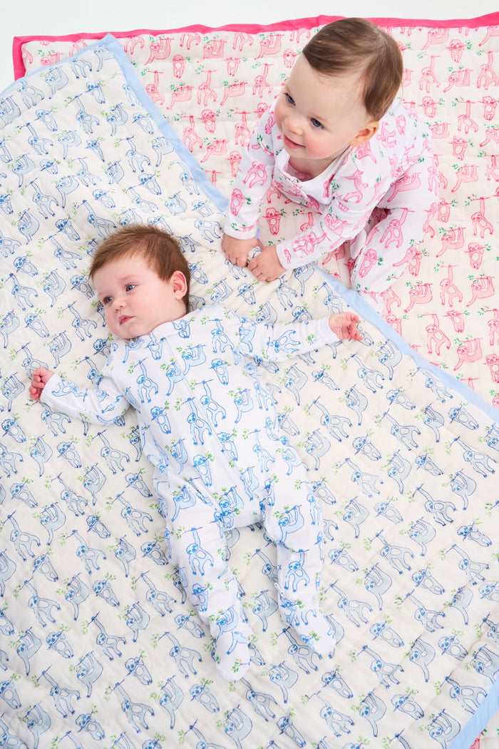 The Roller Rabbit Sleepy Sloth Baby Quilt is a soft cotton, white quilt with a blue border and playful sloth illustrations. Blue and gray sloths hold green leaves, while the inner lining features a light blue striped pattern.