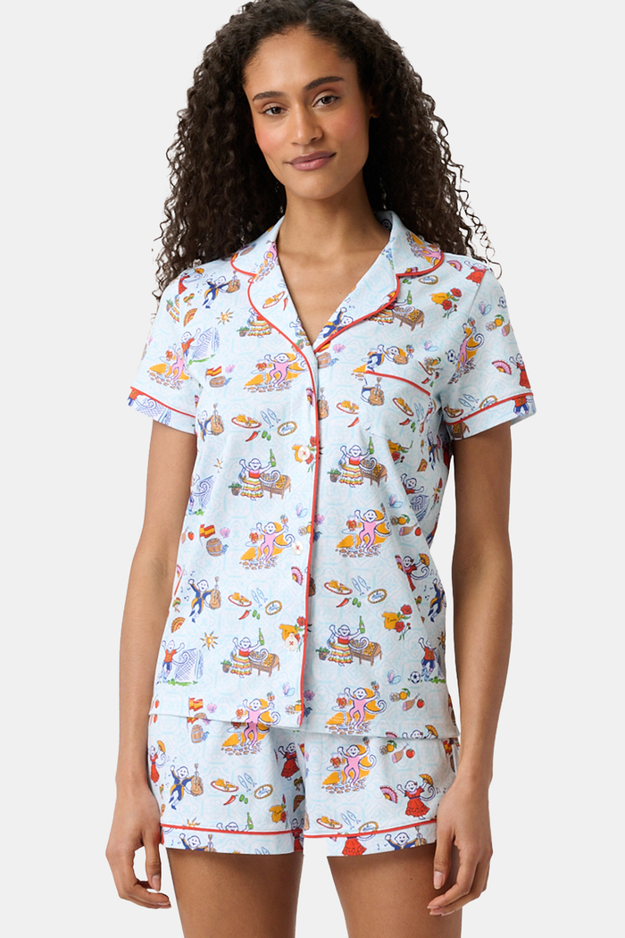 A woman with curly hair smiles in Roller Rabbit Spain Monkey Polo Pajamas—light blue shortie PJs made from 100 Pima cotton with colorful whimsical prints and red piping—standing against a plain white background.