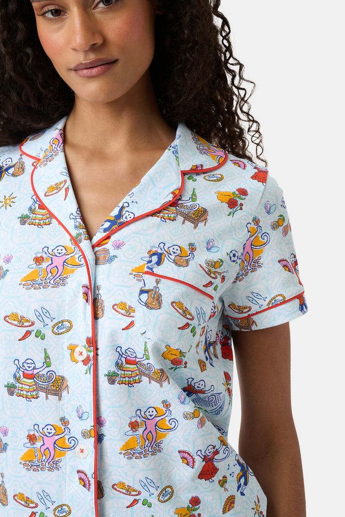 A woman with curly hair smiles in Roller Rabbit Spain Monkey Polo Pajamas—light blue shortie PJs made from 100 Pima cotton with colorful whimsical prints and red piping—standing against a plain white background.