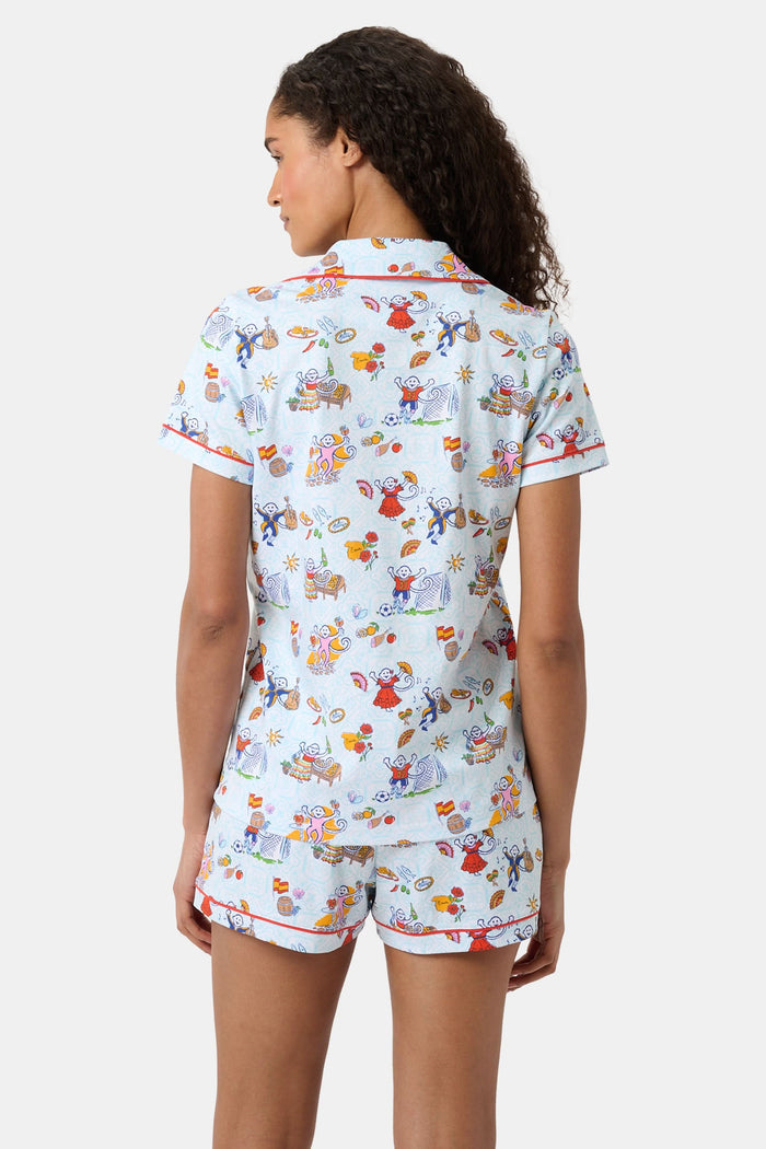 A woman with curly hair smiles in Roller Rabbit Spain Monkey Polo Pajamas—light blue shortie PJs made from 100 Pima cotton with colorful whimsical prints and red piping—standing against a plain white background.