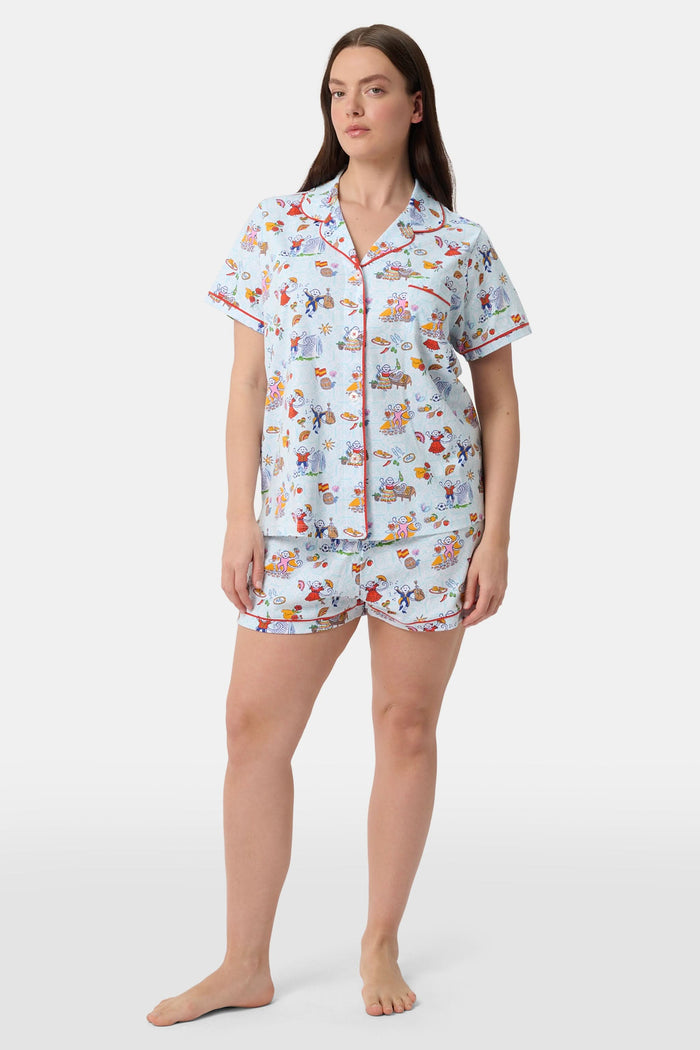 A woman with curly hair smiles in Roller Rabbit Spain Monkey Polo Pajamas—light blue shortie PJs made from 100 Pima cotton with colorful whimsical prints and red piping—standing against a plain white background.