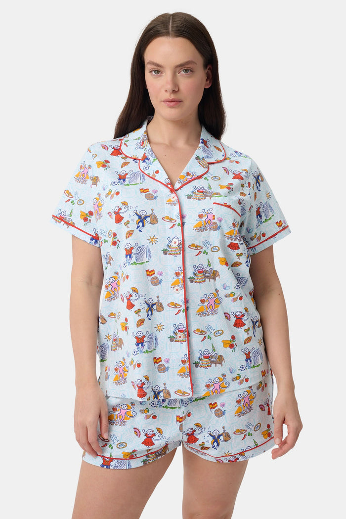 A woman with curly hair smiles in Roller Rabbit Spain Monkey Polo Pajamas—light blue shortie PJs made from 100 Pima cotton with colorful whimsical prints and red piping—standing against a plain white background.