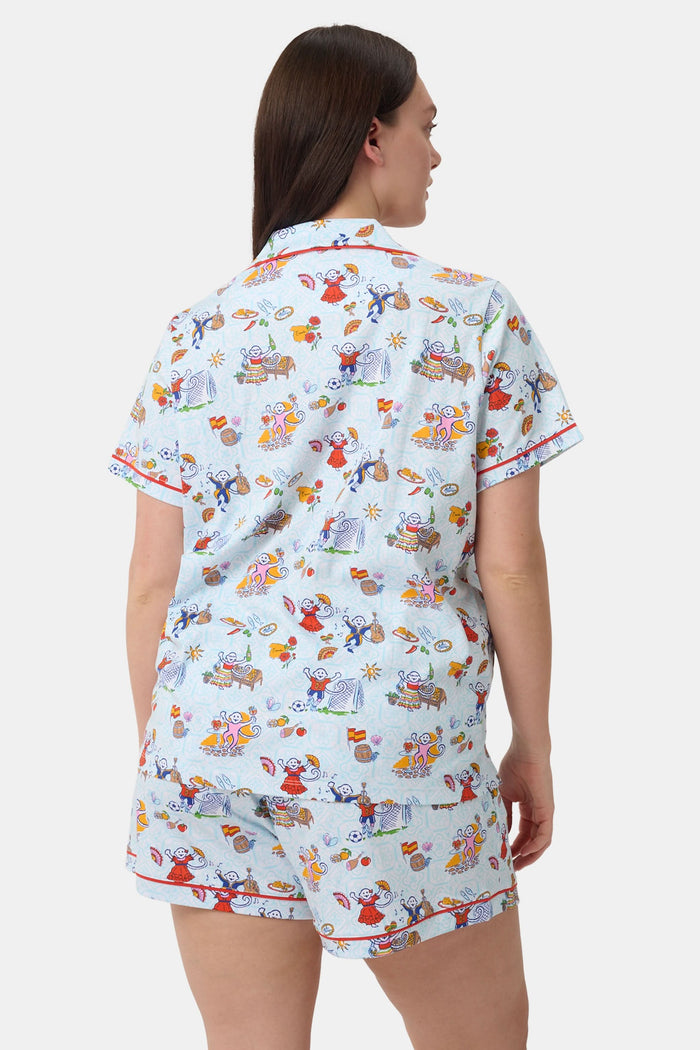 A woman with curly hair smiles in Roller Rabbit Spain Monkey Polo Pajamas—light blue shortie PJs made from 100 Pima cotton with colorful whimsical prints and red piping—standing against a plain white background.