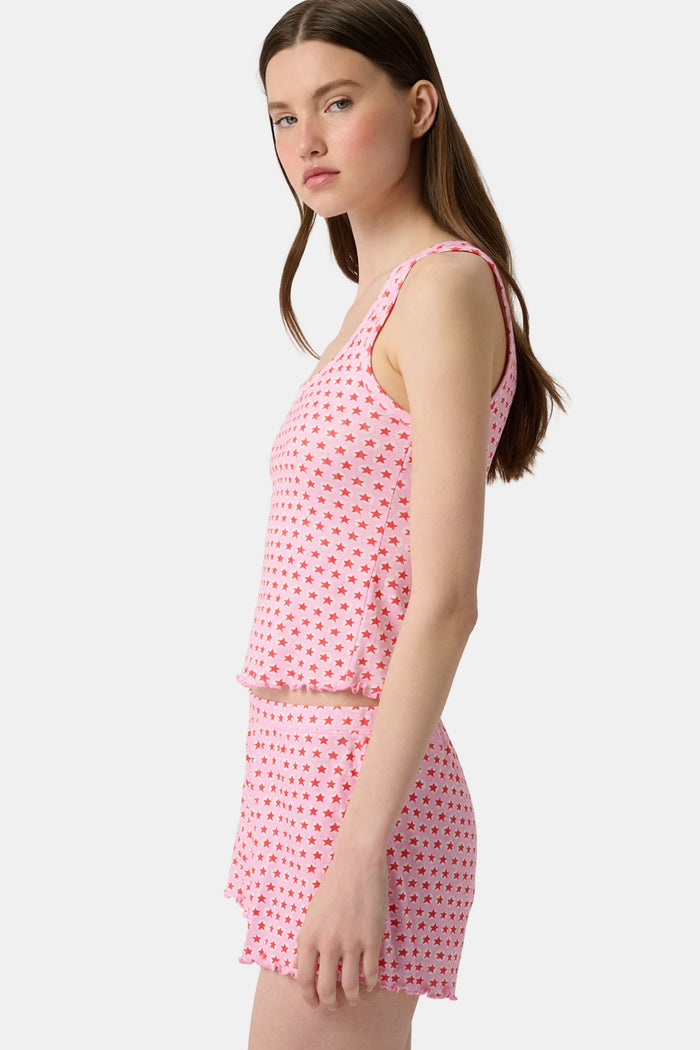 A young woman relaxes with a smile, lying on her back on a boat in the sun. She wears a bubblegum pink Starry Dot Astor Tank by Roller Rabbit and shorts, arms resting behind her head as the water glimmers nearby.