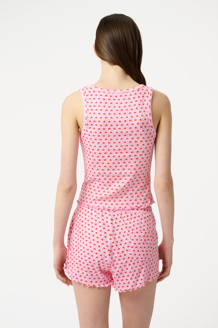 A young woman relaxes with a smile, lying on her back on a boat in the sun. She wears a bubblegum pink Starry Dot Astor Tank by Roller Rabbit and shorts, arms resting behind her head as the water glimmers nearby.