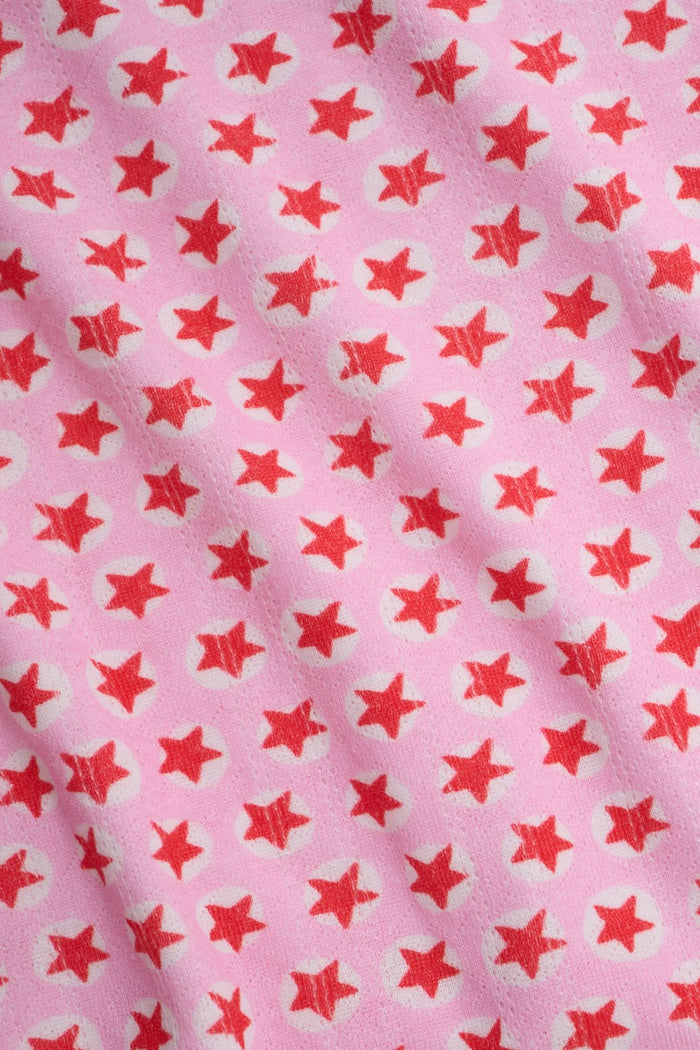 A young woman relaxes with a smile, lying on her back on a boat in the sun. She wears a bubblegum pink Starry Dot Astor Tank by Roller Rabbit and shorts, arms resting behind her head as the water glimmers nearby.