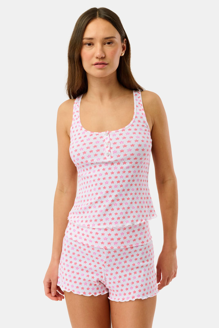 A woman with curly hair stands on a porch, wearing the Roller Rabbit Stars Astor Henley Tank and matching shorts with a pink star pattern. She leans forward on the railing, lush greenery in the background.