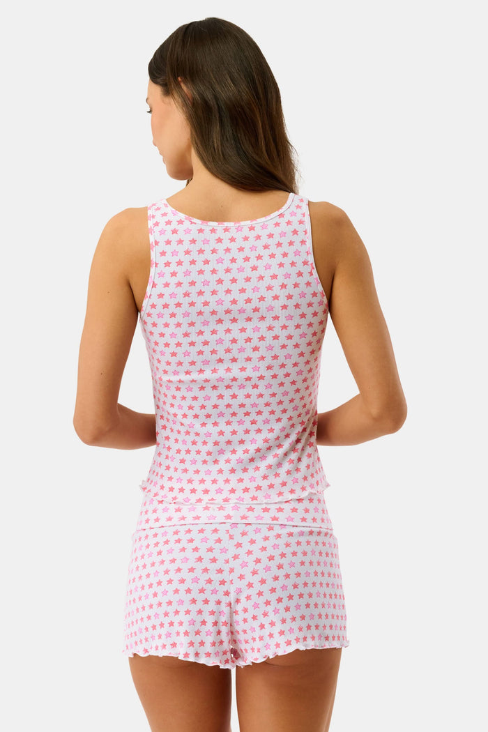 A woman with curly hair stands on a porch, wearing the Roller Rabbit Stars Astor Henley Tank and matching shorts with a pink star pattern. She leans forward on the railing, lush greenery in the background.
