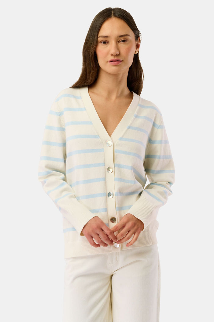 A woman in the Roller Rabbit Stripe Florence Sweater and striped bikini bottoms sits against a white wall, shielding her eyes from the sun with her hand.