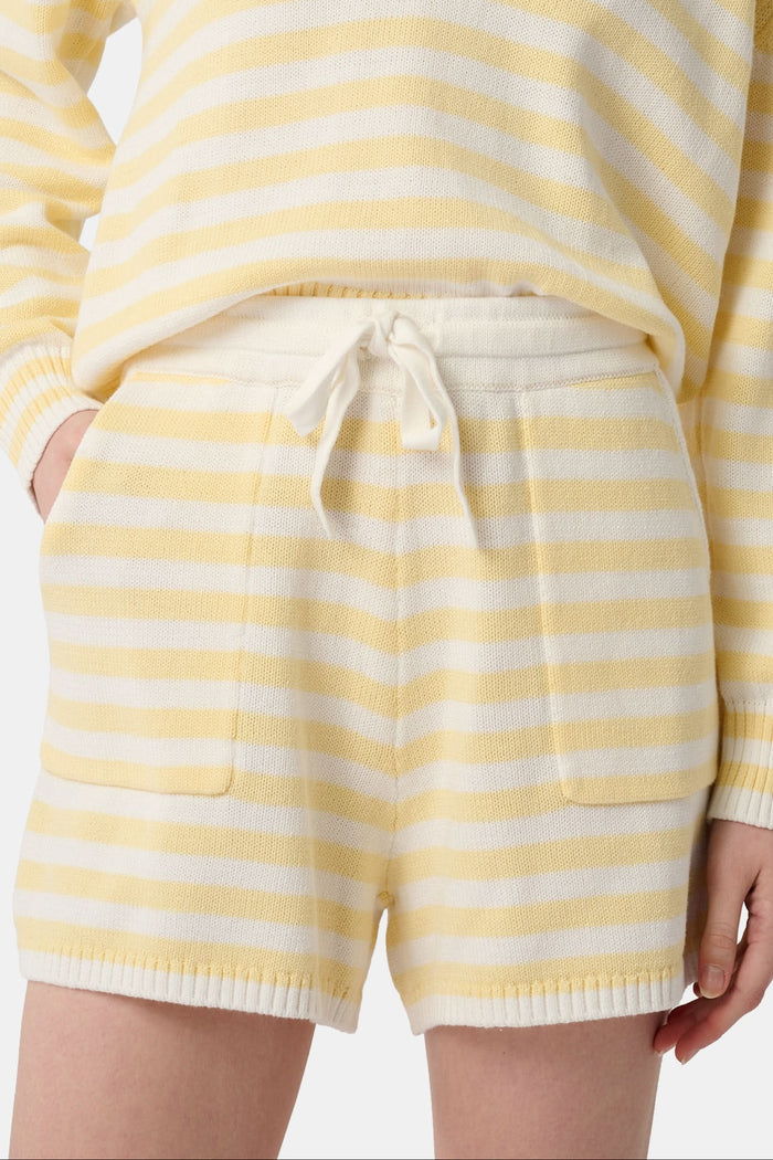 A person wears Roller Rabbit’s Striped Brie Short—a knit yellow and white horizontal striped short with a white drawstring at the elastic waistband and hands tucked in the front pockets.