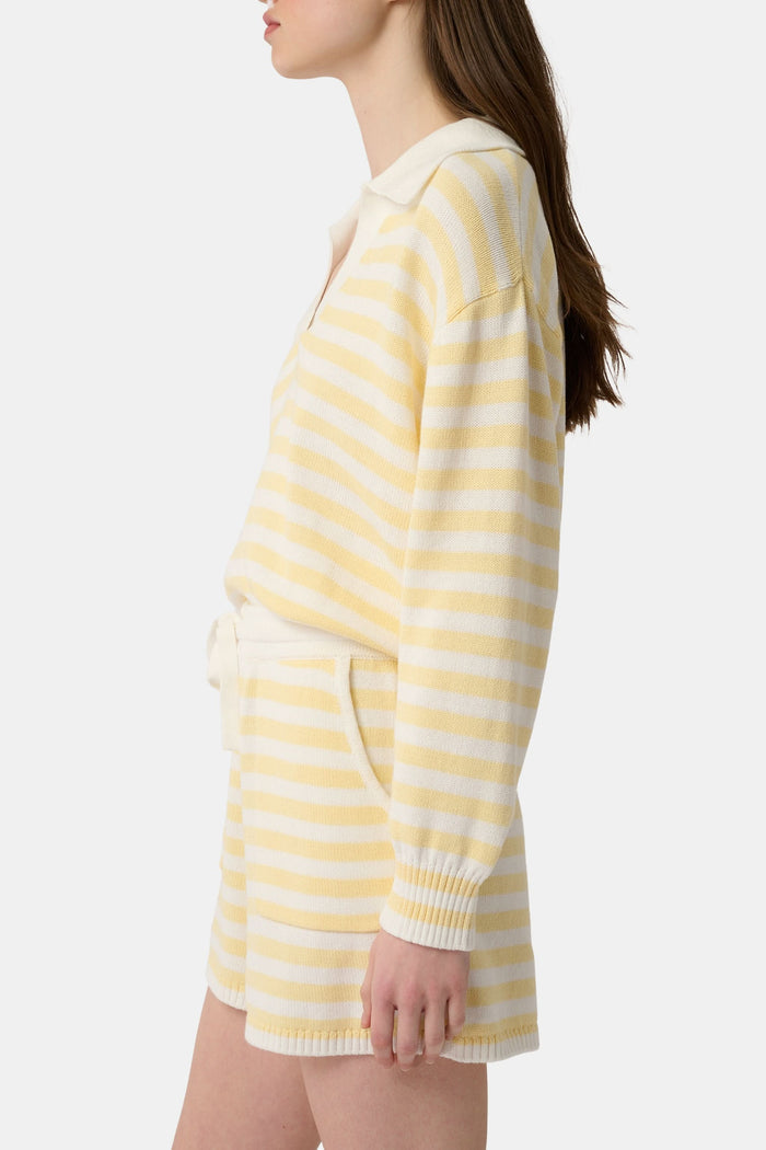 A person wears Roller Rabbit’s Striped Brie Short—a knit yellow and white horizontal striped short with a white drawstring at the elastic waistband and hands tucked in the front pockets.