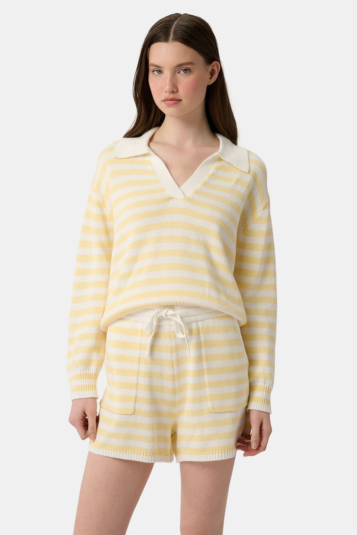 A young woman with light skin and long brown hair wears the Roller Rabbit Striped Rocio Sweater, a pastel yellow, lightweight cotton knit with a wide V-neck and collar. She pairs it with matching drawstring pants and gazes slightly to the side.