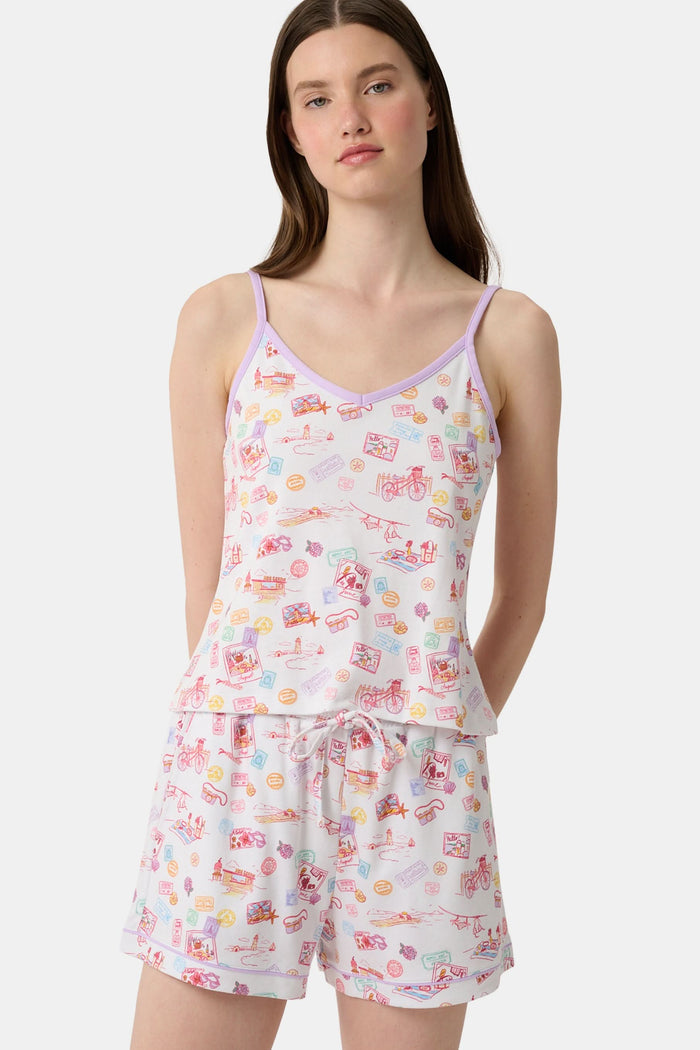 A woman wears the Roller Rabbit Summer Memories Mischa Set, a 100 Pima cotton cami tank and boxer set with a pastel travel icon print on white, standing against a light background.