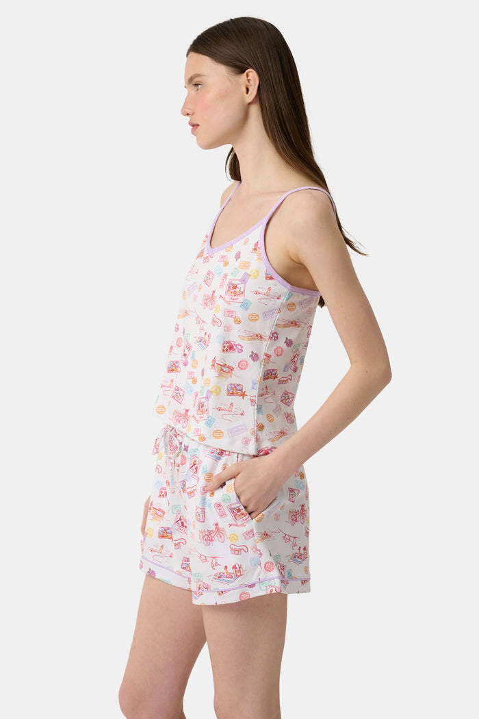 A woman wears the Roller Rabbit Summer Memories Mischa Set, a 100 Pima cotton cami tank and boxer set with a pastel travel icon print on white, standing against a light background.