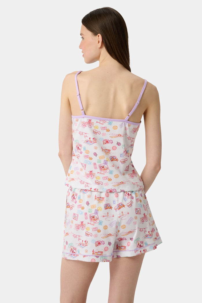 A woman wears the Roller Rabbit Summer Memories Mischa Set, a 100 Pima cotton cami tank and boxer set with a pastel travel icon print on white, standing against a light background.