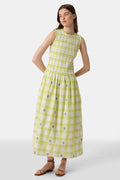 A woman wears the Roller Rabbit Sun Check Blossom Embroidery Emma Dress, a sleeveless green maxi with floral embroidery and smocked bodice, paired with brown sandals. She stands relaxed, looking to the side.