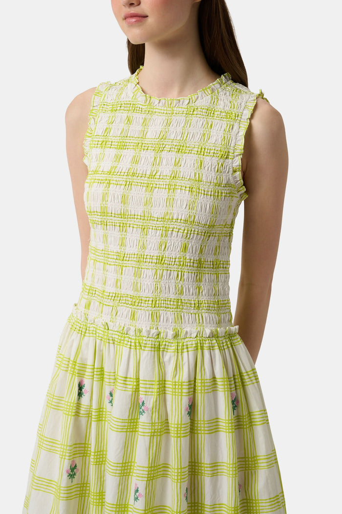 A woman wears the Roller Rabbit Sun Check Blossom Embroidery Emma Dress, a sleeveless green maxi with floral embroidery and smocked bodice, paired with brown sandals. She stands relaxed, looking to the side.