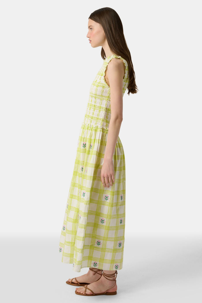 A woman wears the Roller Rabbit Sun Check Blossom Embroidery Emma Dress, a sleeveless green maxi with floral embroidery and smocked bodice, paired with brown sandals. She stands relaxed, looking to the side.