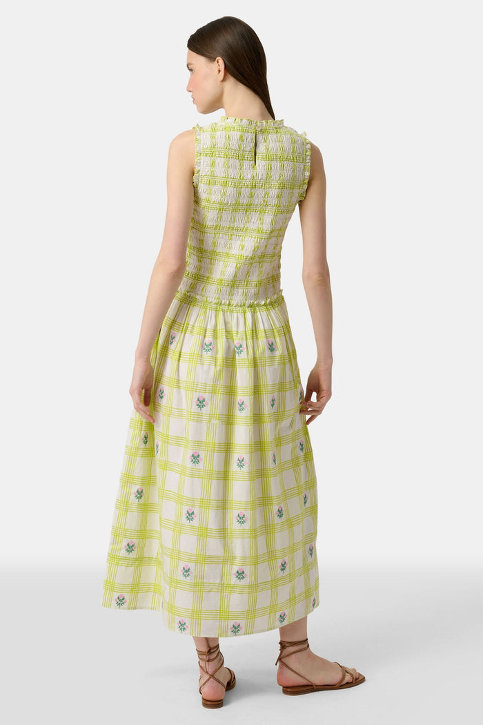 A woman wears the Roller Rabbit Sun Check Blossom Embroidery Emma Dress, a sleeveless green maxi with floral embroidery and smocked bodice, paired with brown sandals. She stands relaxed, looking to the side.