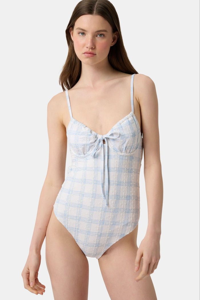 A woman wearing the Roller Rabbit Sun Check Eyelet Aurora One Piece, a light blue and white plaid swimsuit with adjustable straps, stands on a beach with waves and a blue sky in the background.