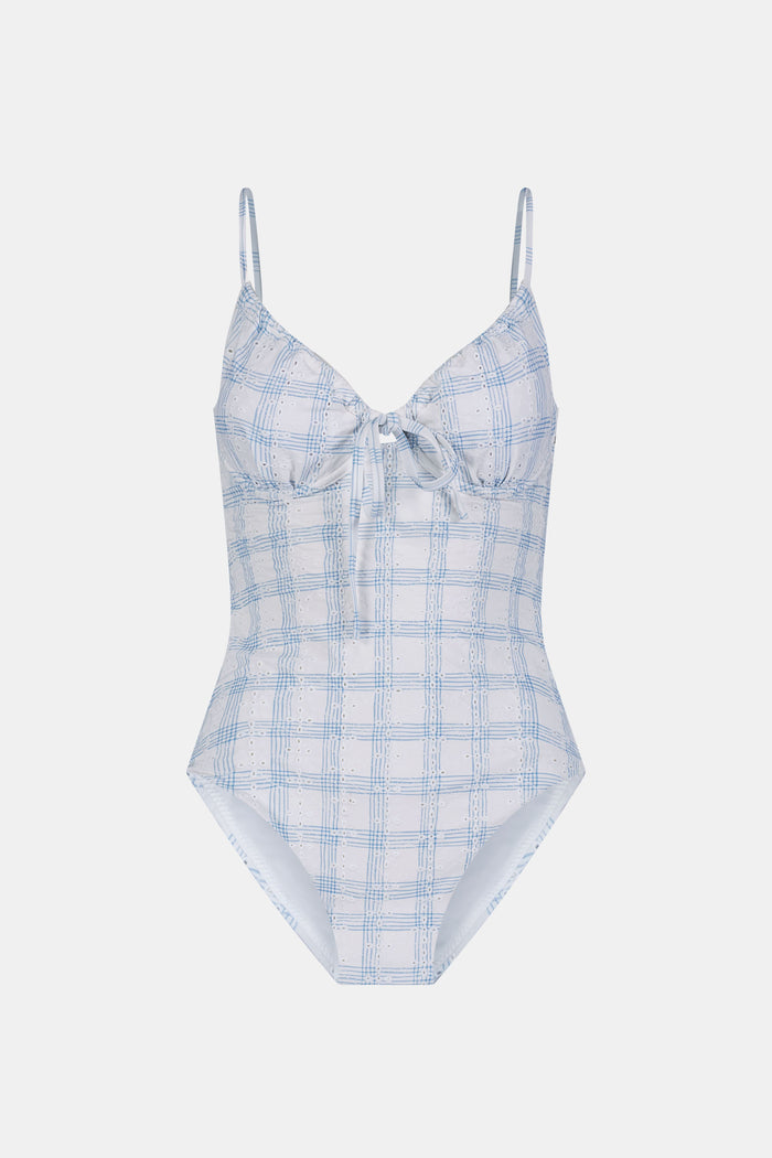 A woman wearing the Roller Rabbit Sun Check Eyelet Aurora One Piece, a light blue and white plaid swimsuit with adjustable straps, stands on a beach with waves and a blue sky in the background.