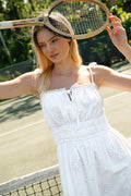 A woman wears the Roller Rabbit Sunny Days Eyelet Viola Dress on a tennis court, holding a vintage racket overhead and smiling slightly. Greenery and part of the net are visible in the background.
