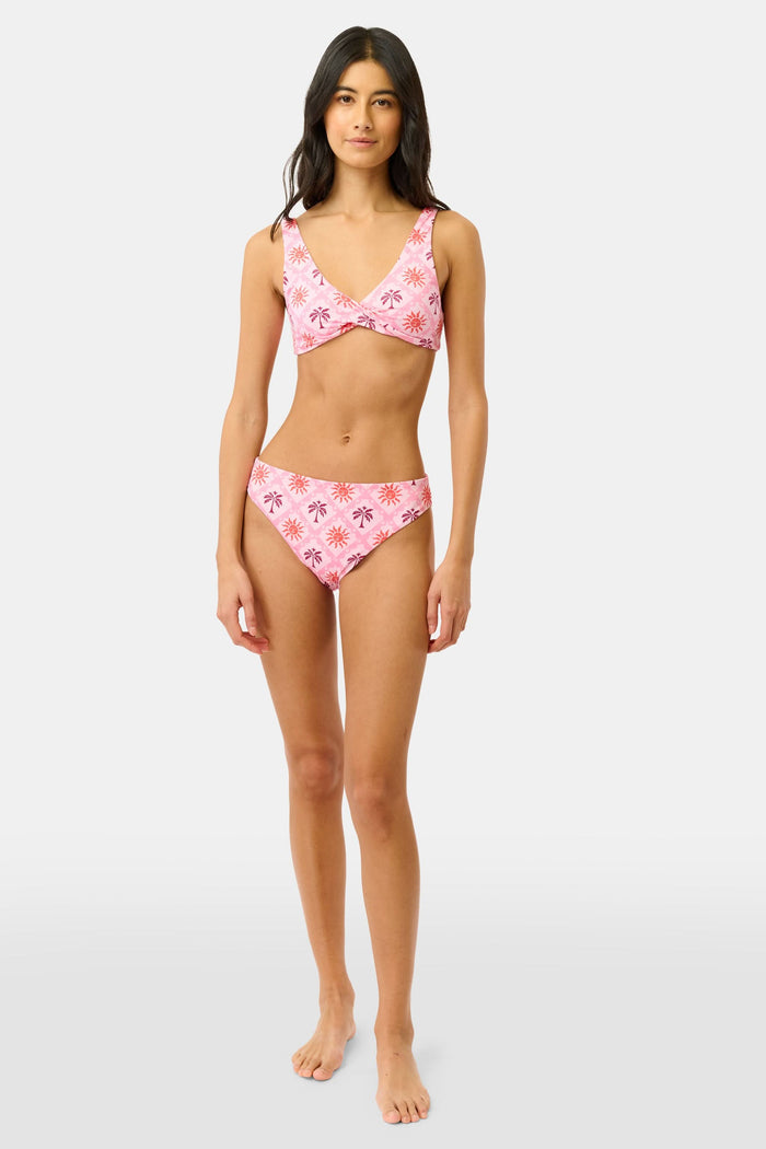 A woman wears the Sunny Days Shelly Bikini Top by Roller Rabbit, a comfy pink and orange wrap bikini featuring red sun and palm tree patterns. She's shown from shoulders to waist, with her face partially out of frame.