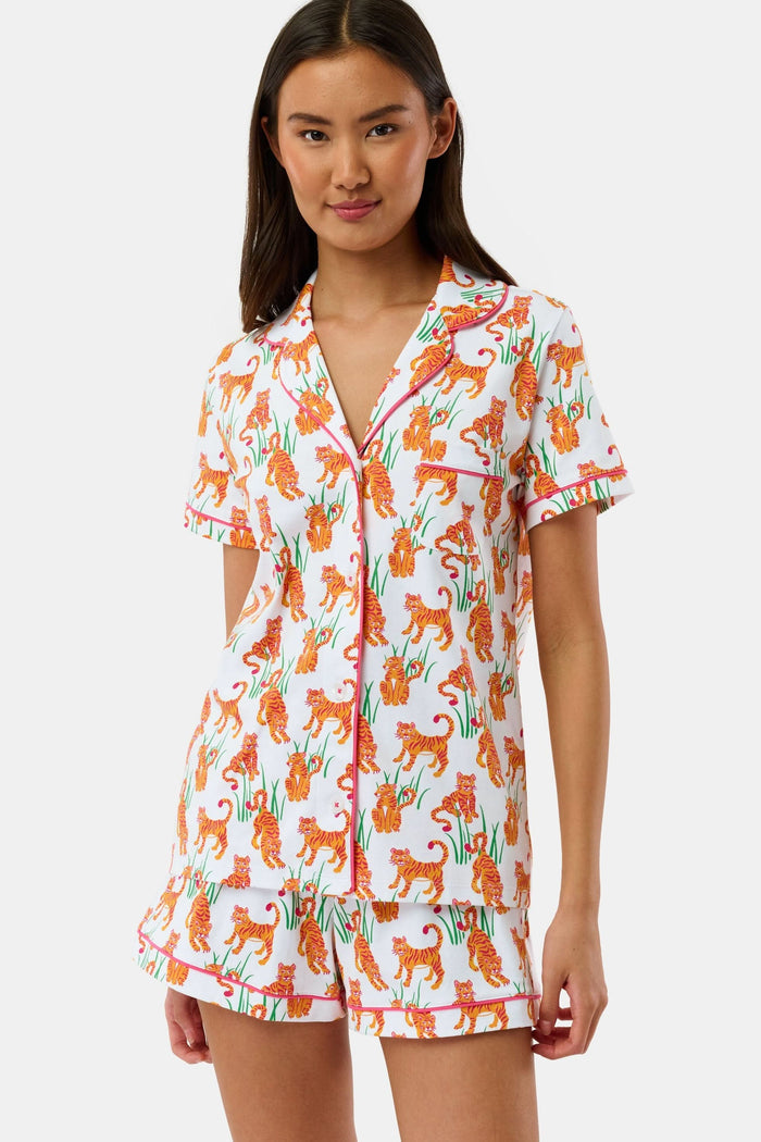 A person wears Roller Rabbit's Teddy Tiger Polo Pajamas, a matching short-sleeve set in 100 Pima Cotton, featuring a green plant and tiger print on white. They stand and smile slightly.