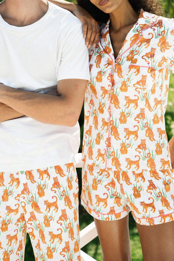 A person wears Roller Rabbit's Teddy Tiger Polo Pajamas, a matching short-sleeve set in 100 Pima Cotton, featuring a green plant and tiger print on white. They stand and smile slightly.