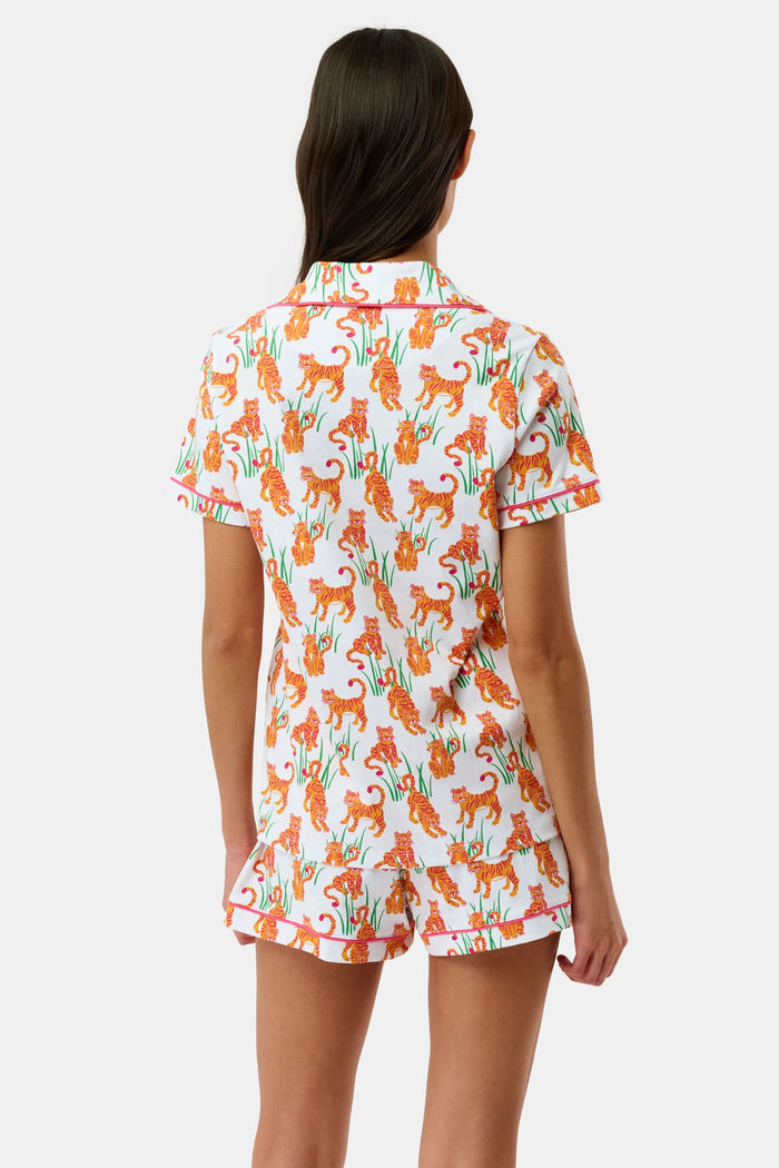 A person wears Roller Rabbit's Teddy Tiger Polo Pajamas, a matching short-sleeve set in 100 Pima Cotton, featuring a green plant and tiger print on white. They stand and smile slightly.