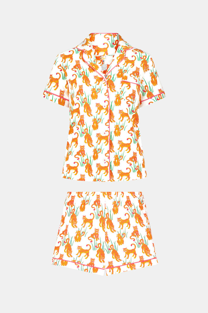 A person wears Roller Rabbit's Teddy Tiger Polo Pajamas, a matching short-sleeve set in 100 Pima Cotton, featuring a green plant and tiger print on white. They stand and smile slightly.