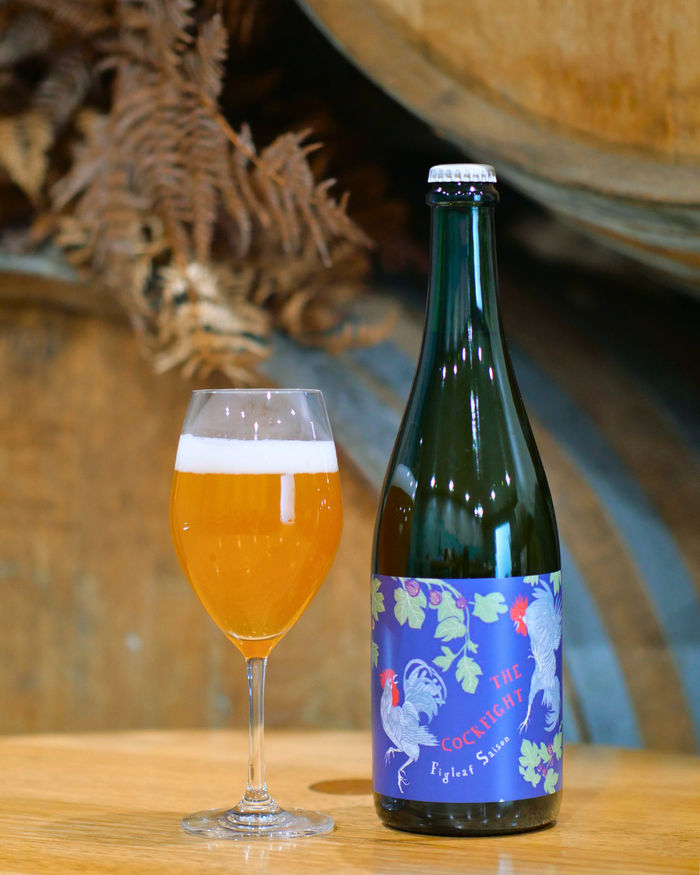 Bottle of The Cockfight, Sailors Grave barrel aged beer with a decorative label next to a glass of beer on a wooden surface.