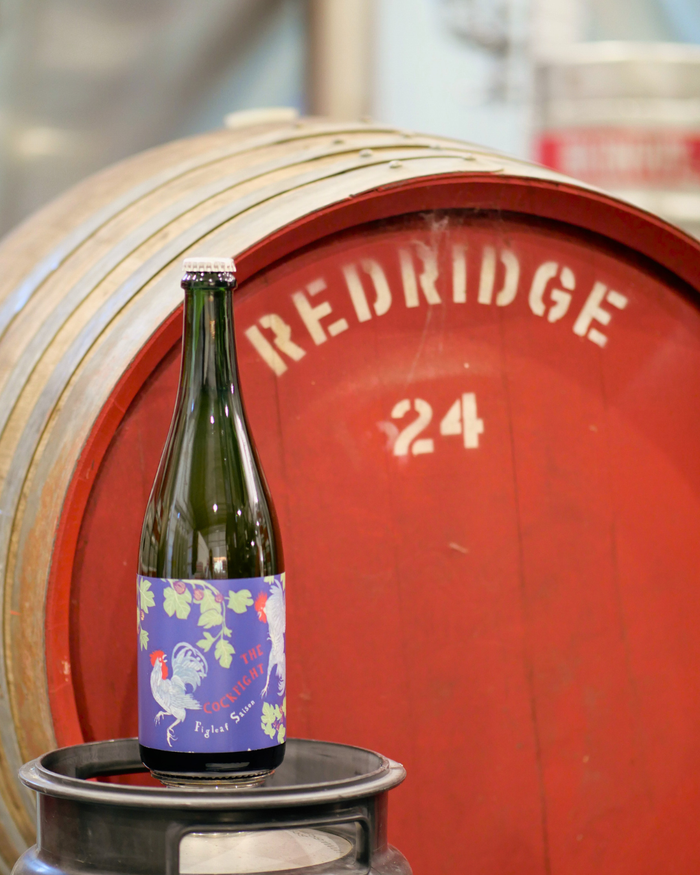 Bottle of The Cockfight beer on a keg in front of a red barrel labeled 'Redridge 24'.