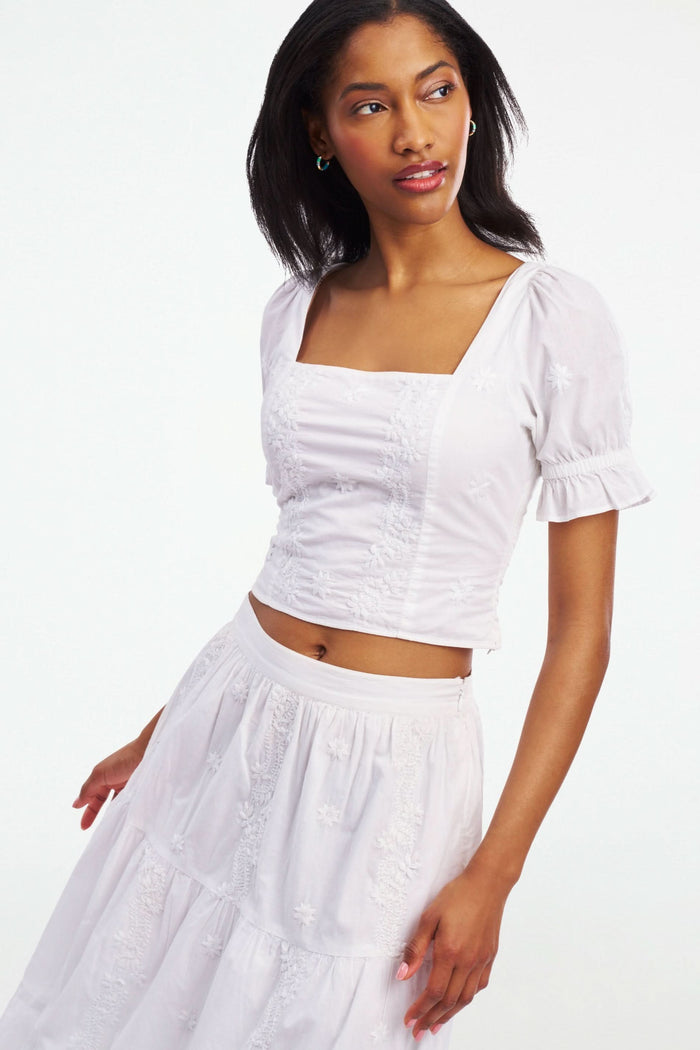 A woman wearing the Roller Rabbit Vanna Embroidery Kayla Top, a white blouse with short sleeves and a square neckline, paired with a matching high-waisted skirt, stands against a plain white background.