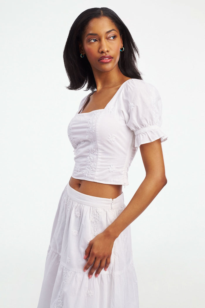 A woman wearing the Roller Rabbit Vanna Embroidery Kayla Top, a white blouse with short sleeves and a square neckline, paired with a matching high-waisted skirt, stands against a plain white background.