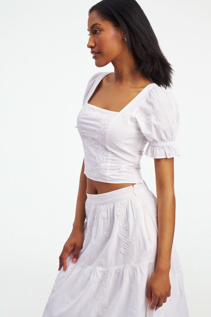 A woman wearing the Roller Rabbit Vanna Embroidery Kayla Top, a white blouse with short sleeves and a square neckline, paired with a matching high-waisted skirt, stands against a plain white background.