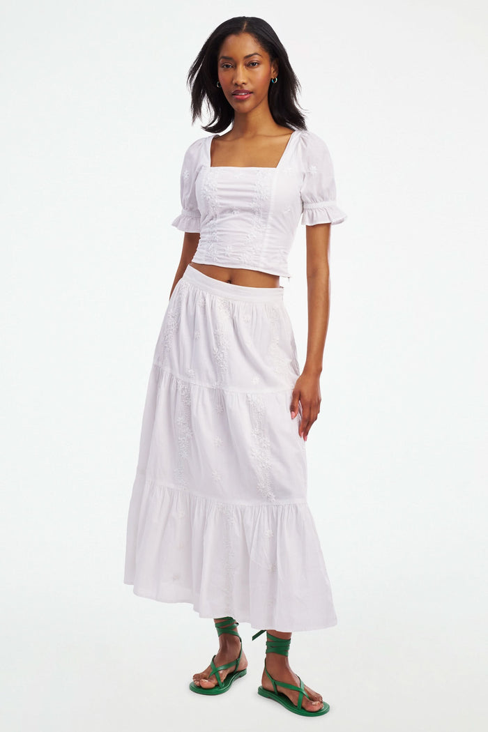 A woman wearing the Roller Rabbit Vanna Embroidery Kayla Top, a white blouse with short sleeves and a square neckline, paired with a matching high-waisted skirt, stands against a plain white background.