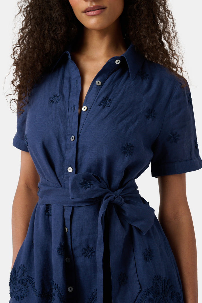 A woman models the Roller Rabbit Vanna Embroidery Margaret Dress—a short-sleeve navy linen shirtdress with a belted waist—standing against a plain light background.