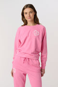 A person wearing the Roller Rabbit Vintage Classic Crewneck in pink, paired with matching pink sweatpants, stands against a plain background and looks at the camera with a slight smile. The outfit features small heart designs.