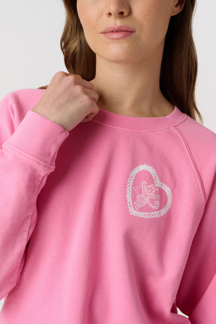 A person wearing the Roller Rabbit Vintage Classic Crewneck in pink, paired with matching pink sweatpants, stands against a plain background and looks at the camera with a slight smile. The outfit features small heart designs.