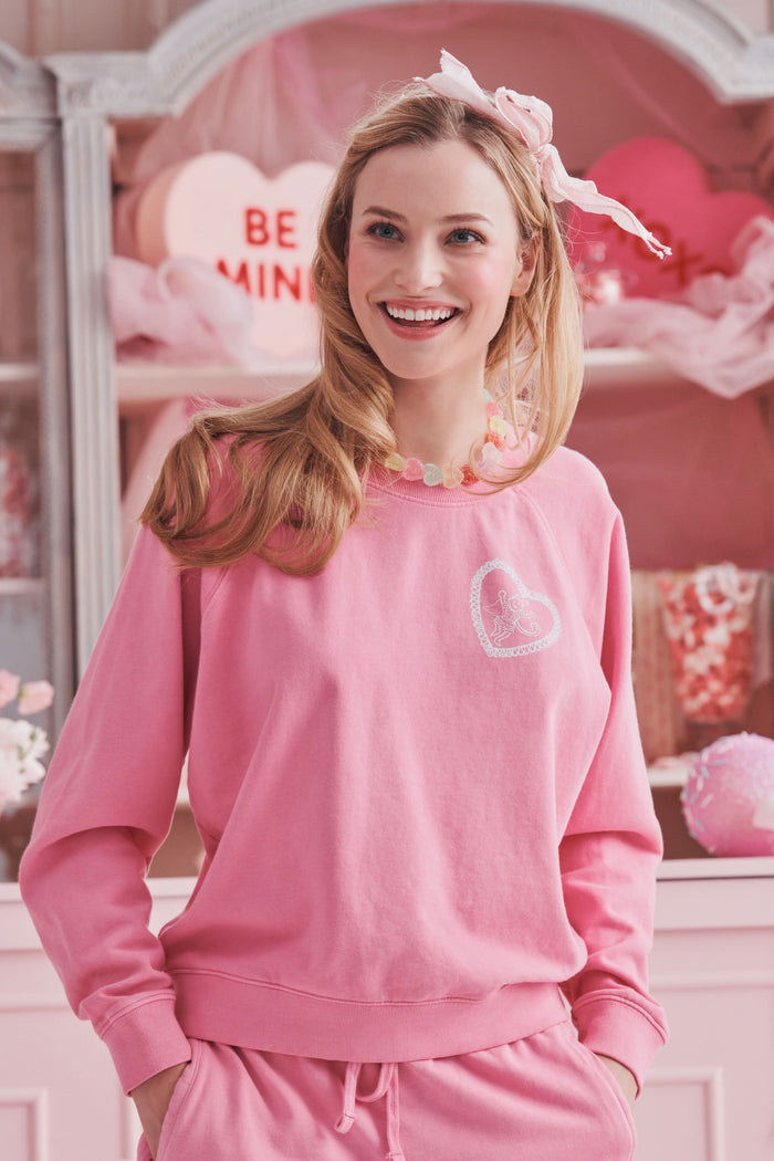 A person wearing the Roller Rabbit Vintage Classic Crewneck in pink, paired with matching pink sweatpants, stands against a plain background and looks at the camera with a slight smile. The outfit features small heart designs.