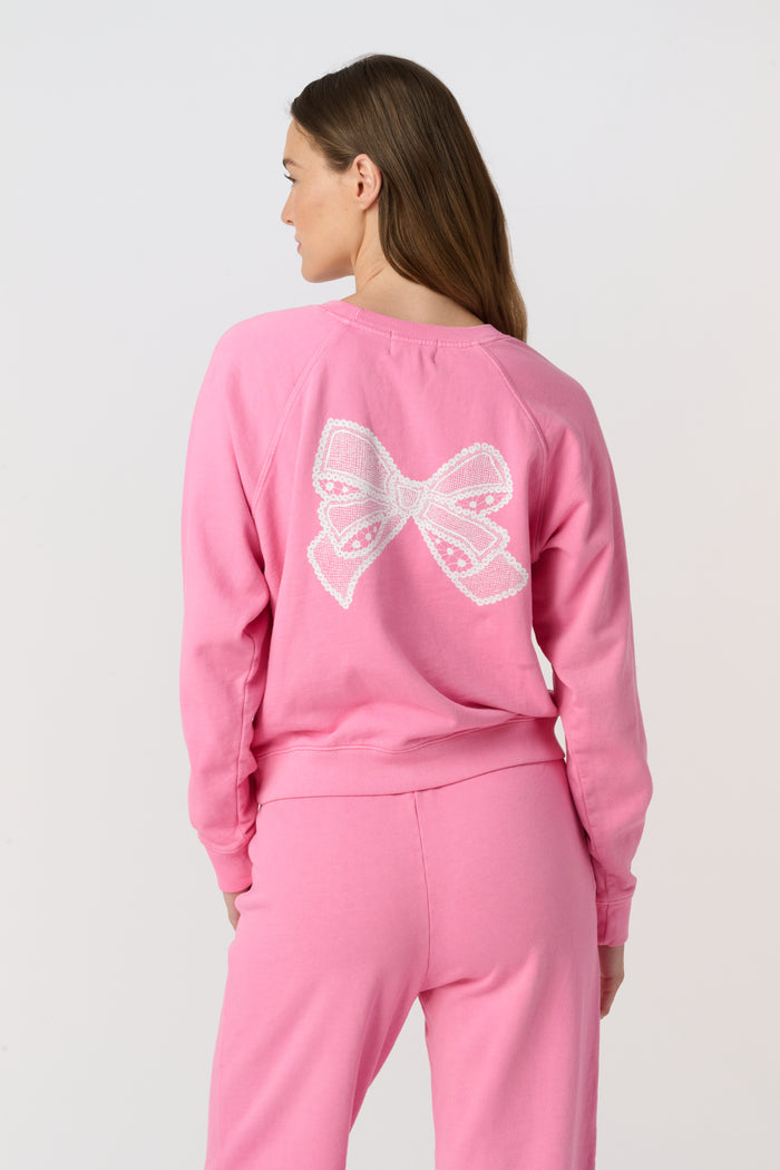 A person wearing the Roller Rabbit Vintage Classic Crewneck in pink, paired with matching pink sweatpants, stands against a plain background and looks at the camera with a slight smile. The outfit features small heart designs.