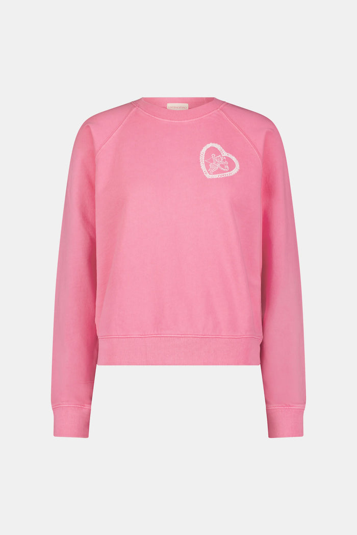 A person wearing the Roller Rabbit Vintage Classic Crewneck in pink, paired with matching pink sweatpants, stands against a plain background and looks at the camera with a slight smile. The outfit features small heart designs.