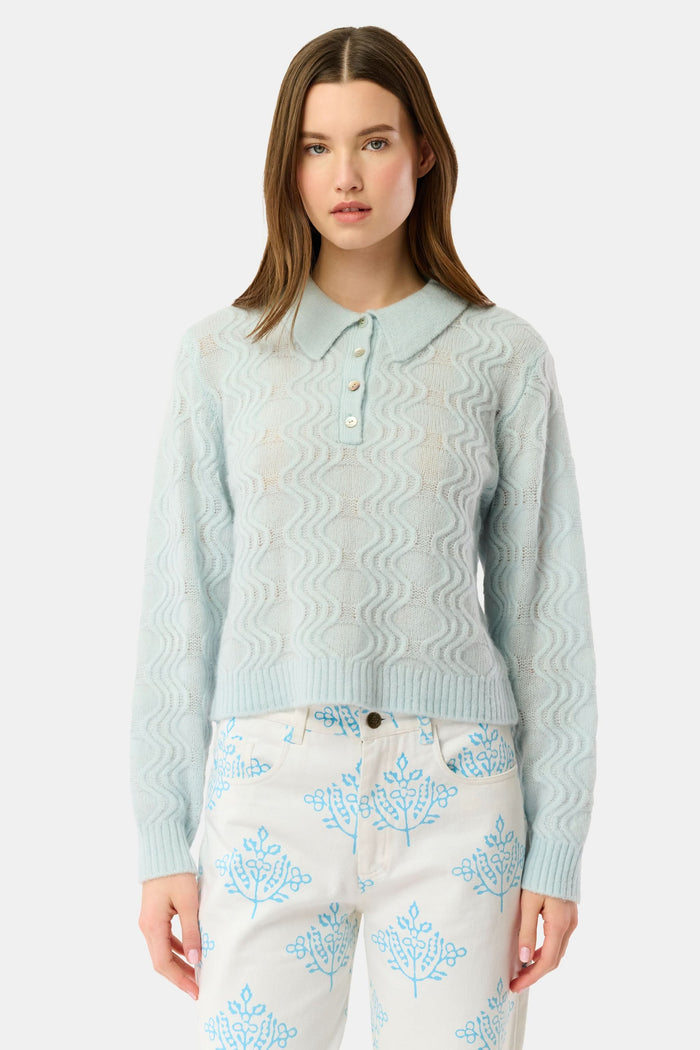 A woman with straight brown hair wears the Roller Rabbit Wave Knit Ava Sweater, a light blue collared, button-up style, paired with white pants with a blue floral print. She stands against a plain light background.