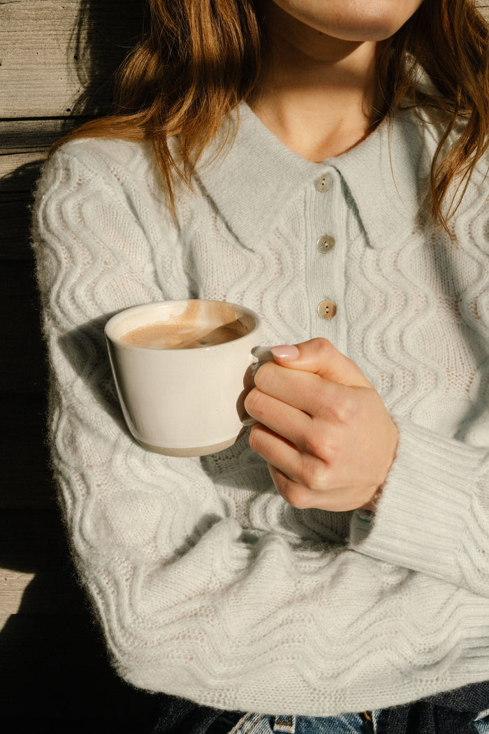 A woman with straight brown hair wears the Roller Rabbit Wave Knit Ava Sweater, a light blue collared, button-up style, paired with white pants with a blue floral print. She stands against a plain light background.