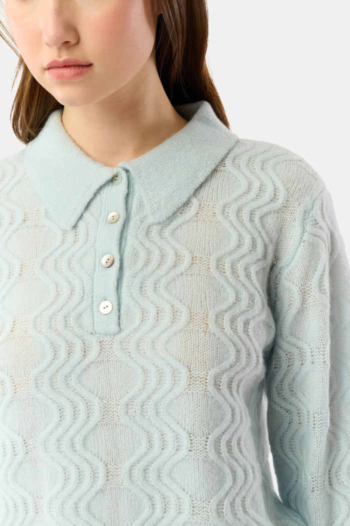 A woman with straight brown hair wears the Roller Rabbit Wave Knit Ava Sweater, a light blue collared, button-up style, paired with white pants with a blue floral print. She stands against a plain light background.