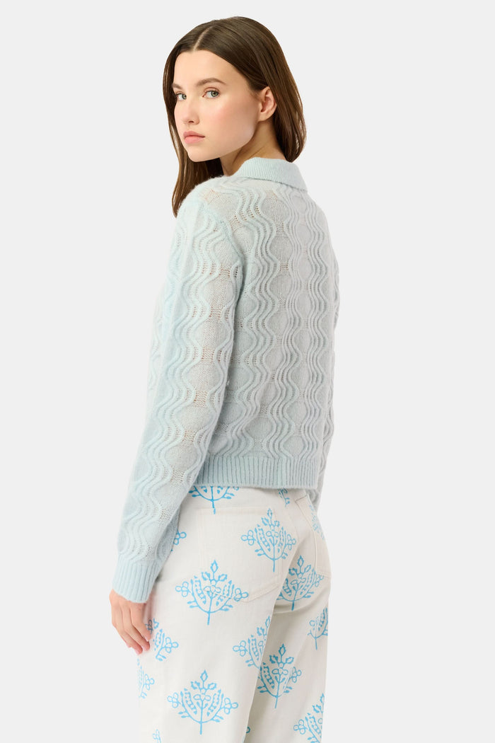 A woman with straight brown hair wears the Roller Rabbit Wave Knit Ava Sweater, a light blue collared, button-up style, paired with white pants with a blue floral print. She stands against a plain light background.